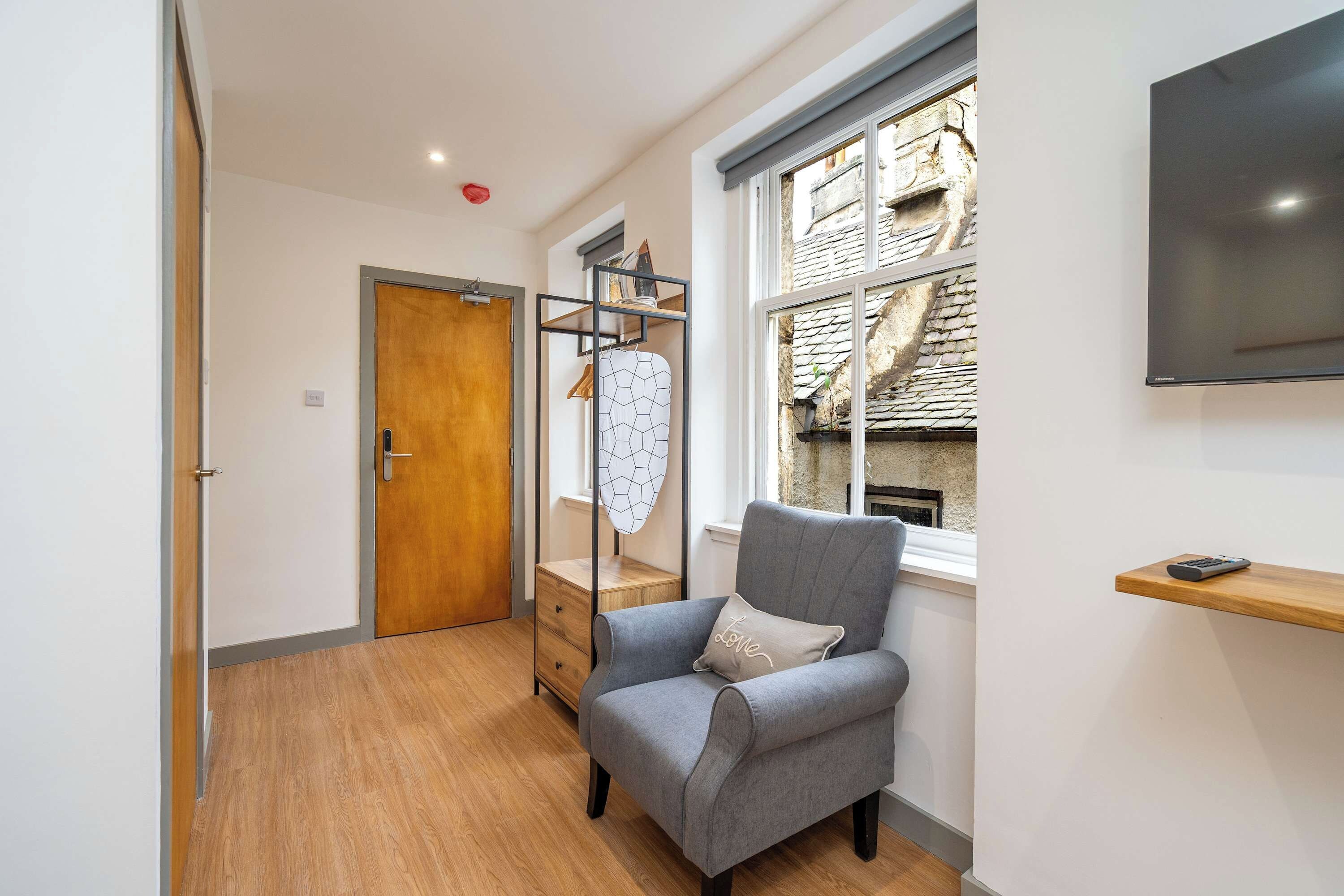 A modern room with a grey armchair, wooden floor, a wooden door, a window with a view of a stone building, and a flat-screen TV mounted on the wall.