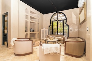 Una habitación con zona de estar, que incluye dos sillones y un sofá, una mesita con una bandeja de objetos y un ventanal decorativo en arco.