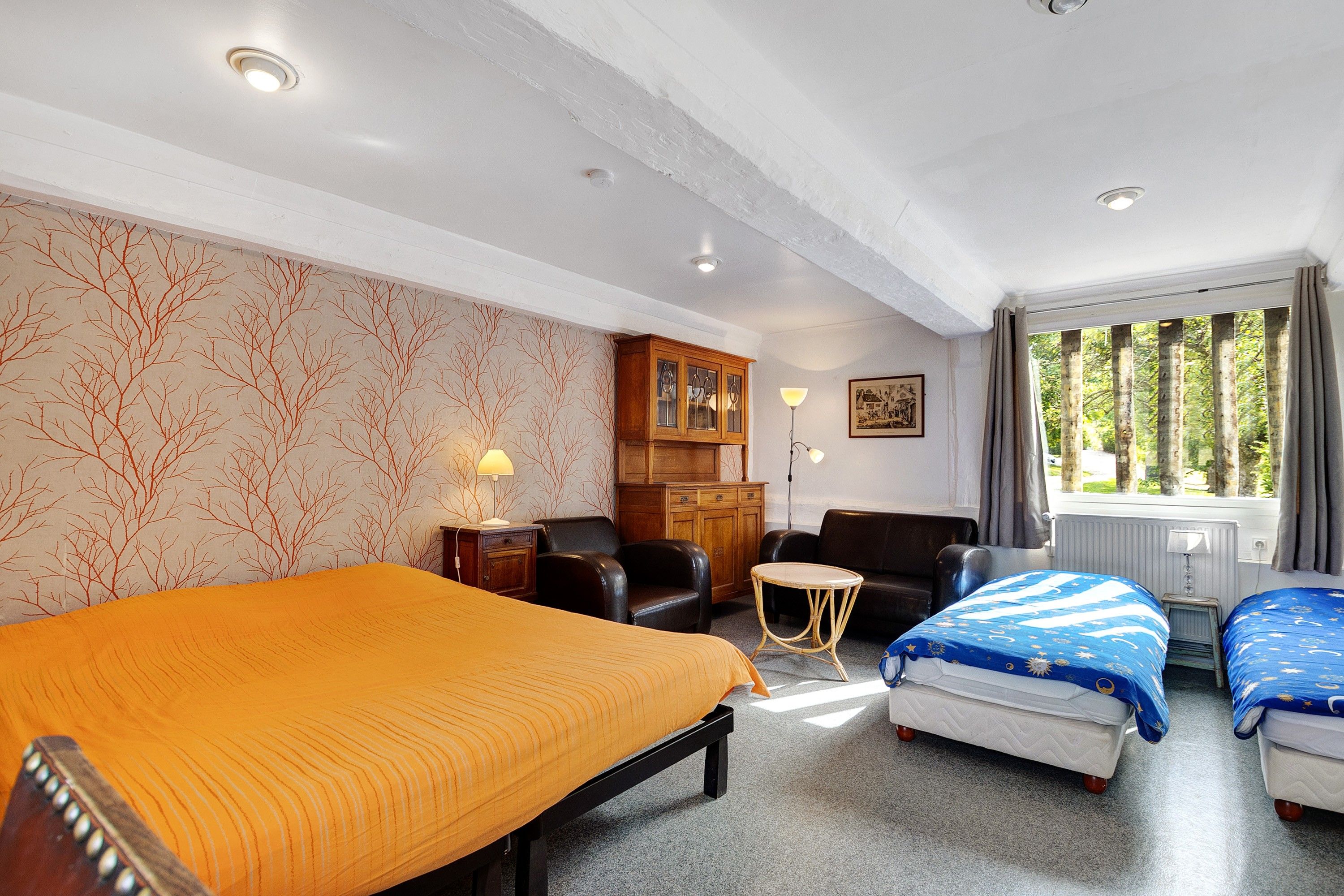 A hotel room with a large bed, two smaller beds, a desk, a chair, a cabinet, and a window with a view of trees.