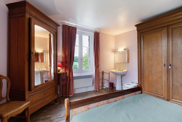 A bedroom with a wooden wardrobe, a bedside table, a mirror, a window with curtains, a bedside lamp, and a bed.