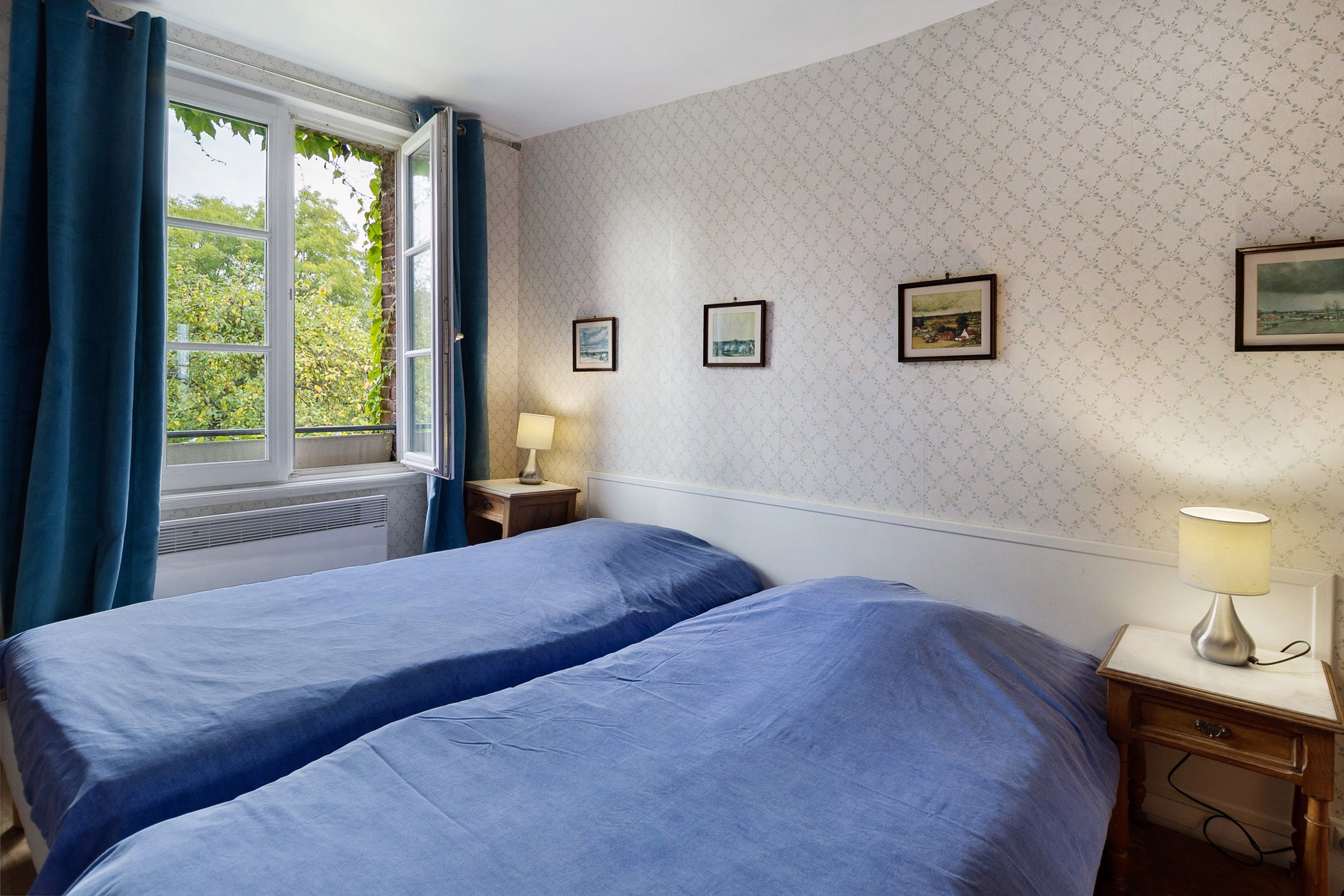 Een kamer met twee bedden, een raam met gordijnen, een nachtkastje met een lamp en drie ingelijste foto's aan de muur.
