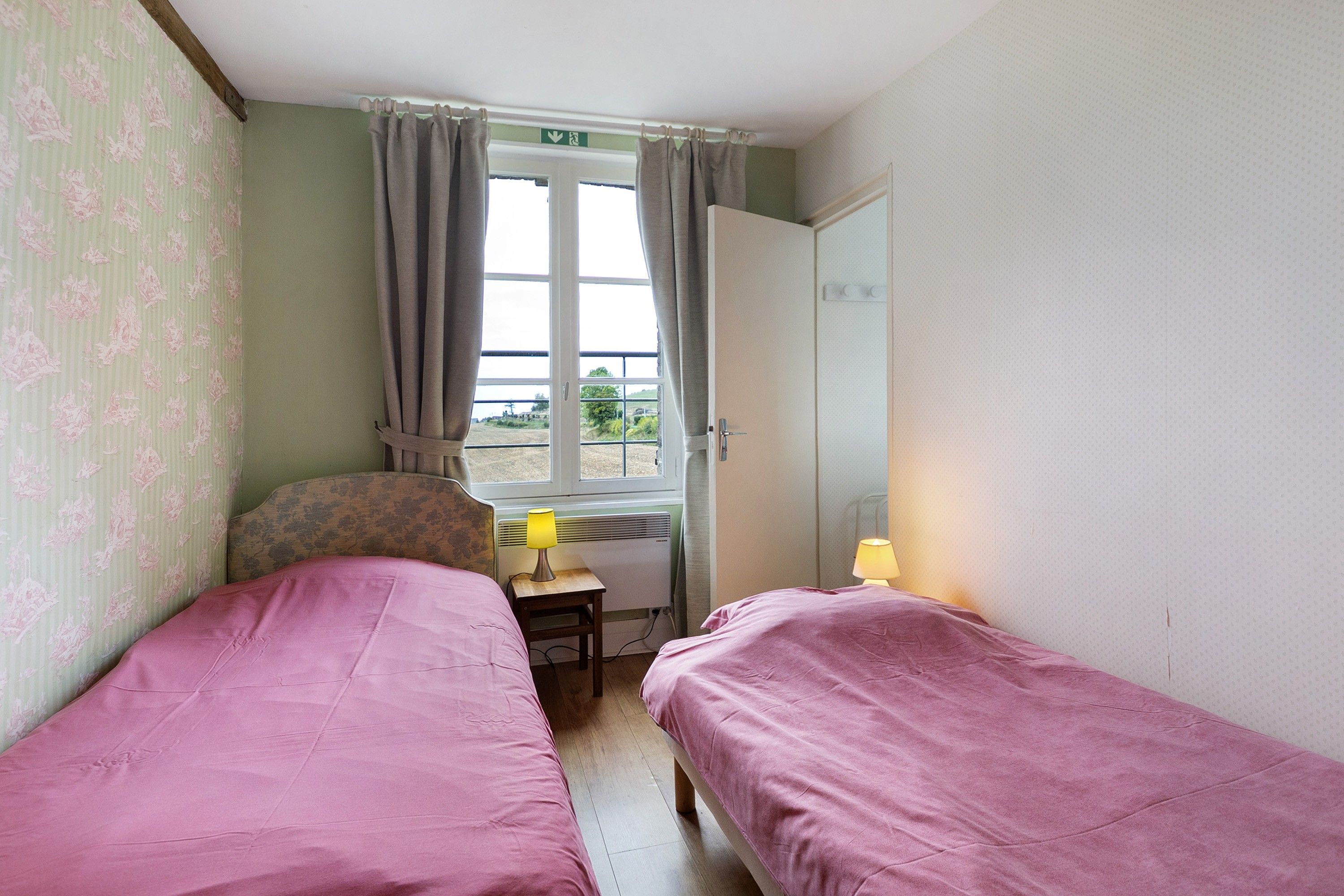 A room with two beds, a window with curtains, a door, and a small table with a lamp.