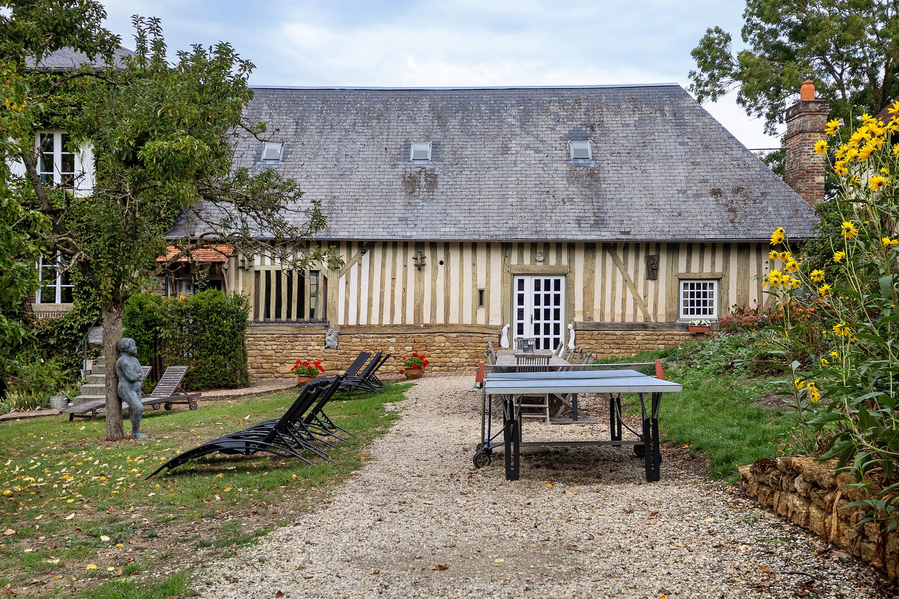 A traditional building with a stone facade, a gravel path leading to a table and chairs, and a statue in the foreground.