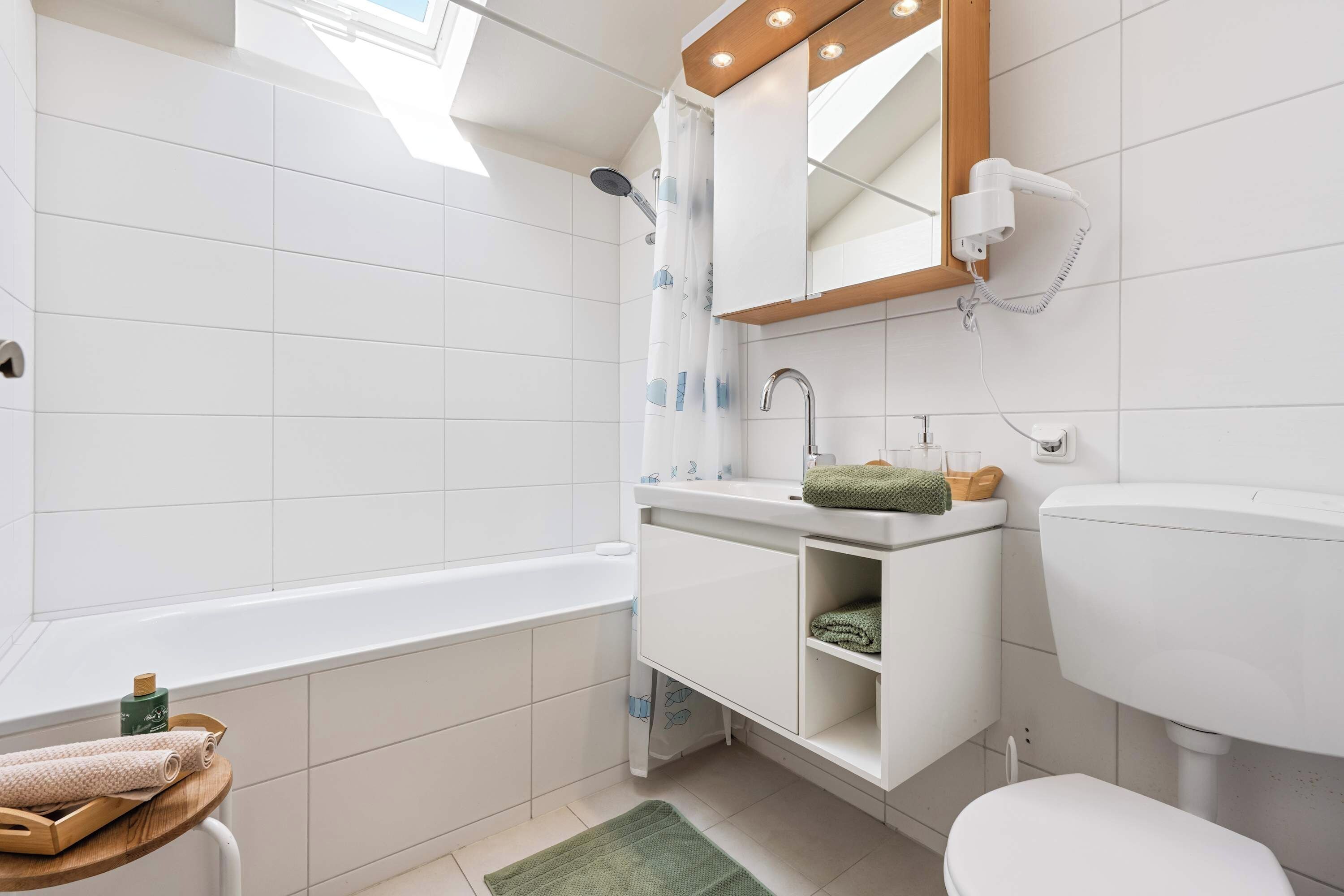 A modern bathroom with a bathtub, a sink, and a toilet. There is a wooden shelf with towels and a green mat on the floor.