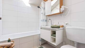 A modern bathroom with a bathtub, a sink, and a toilet. There is a wooden shelf with towels and a green mat on the floor.