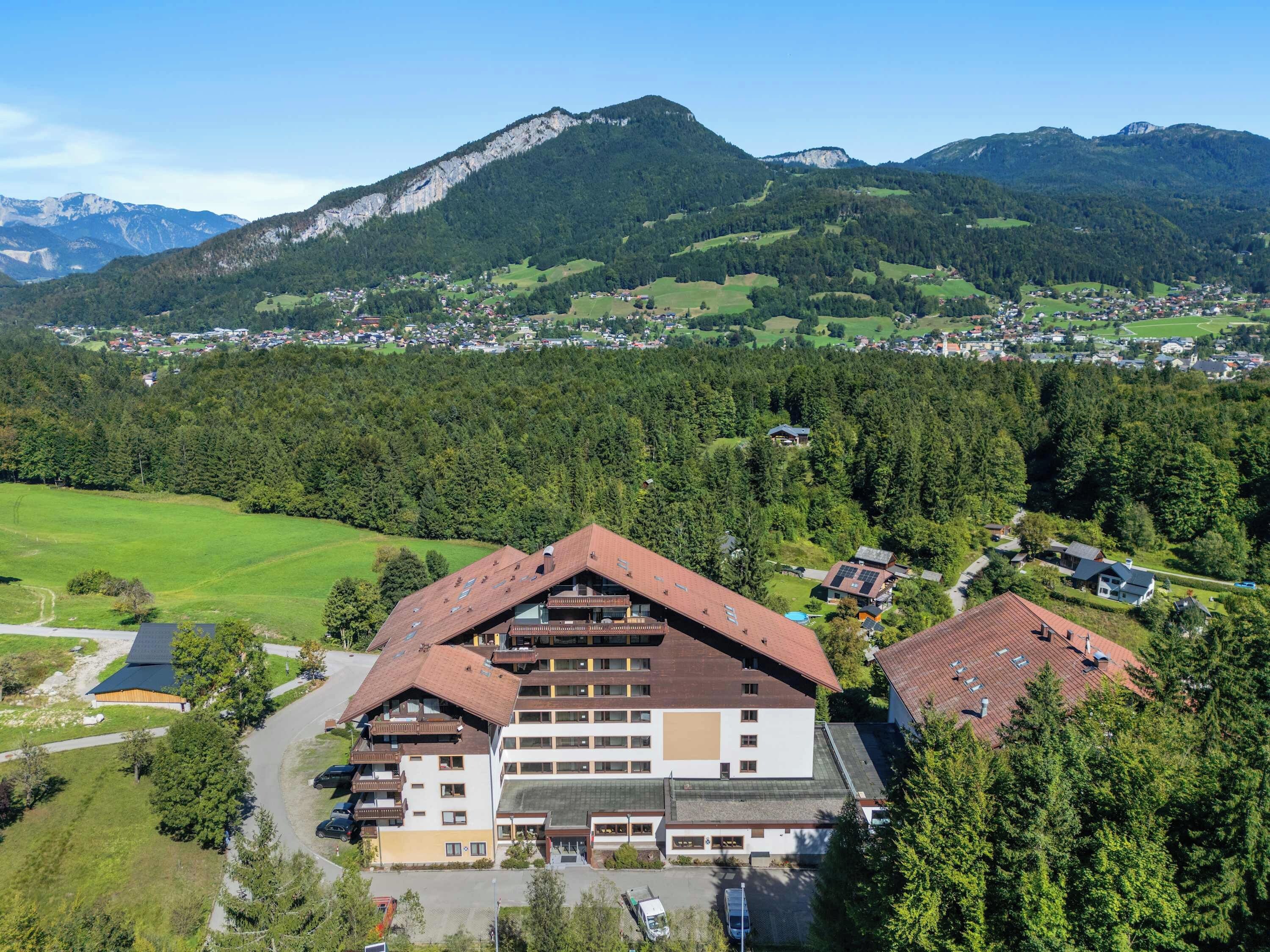 A multi-story building with a sloped roof, surrounded by a forested area and mountains in the background.