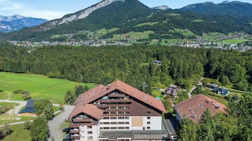 A multi-story building with a sloped roof, surrounded by a forested area and mountains in the background.