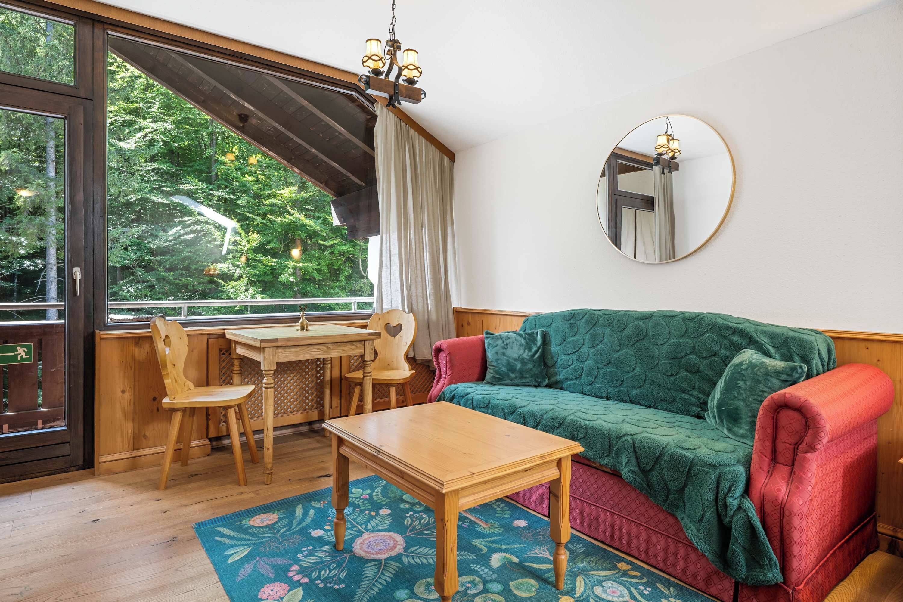Une pièce chaleureuse avec une table en bois, des chaises, un canapé vert, un miroir et une vue sur la verdure à l’extérieur.