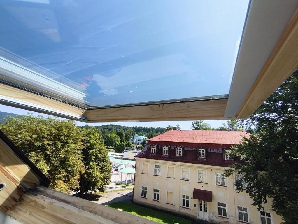 Blick durch ein Fenster zu einem klaren Himmel, einem Gebäude mit rotem Dach und Bäumen im Hintergrund.