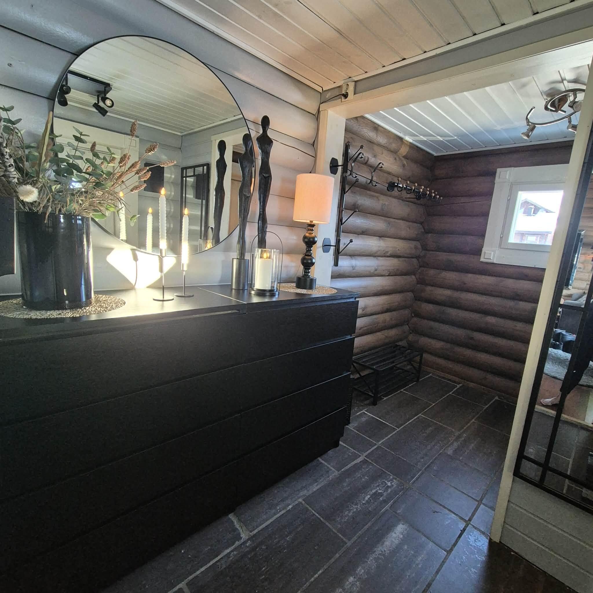 Interior de una cabaña de troncos con un tocador de madera oscura, un espejo y una ventana con vista a una casa.