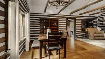 A rustic interior with a wooden dining table, chairs, a bookshelf, and a cozy seating area with a sofa and armchair.