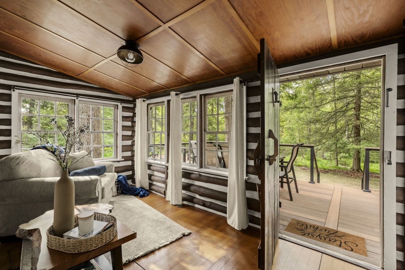 Ein Holzblockhaus-Interieur mit einer Couch, einem Tisch mit einer Vase und Blick ins Freie durch große Fenster.