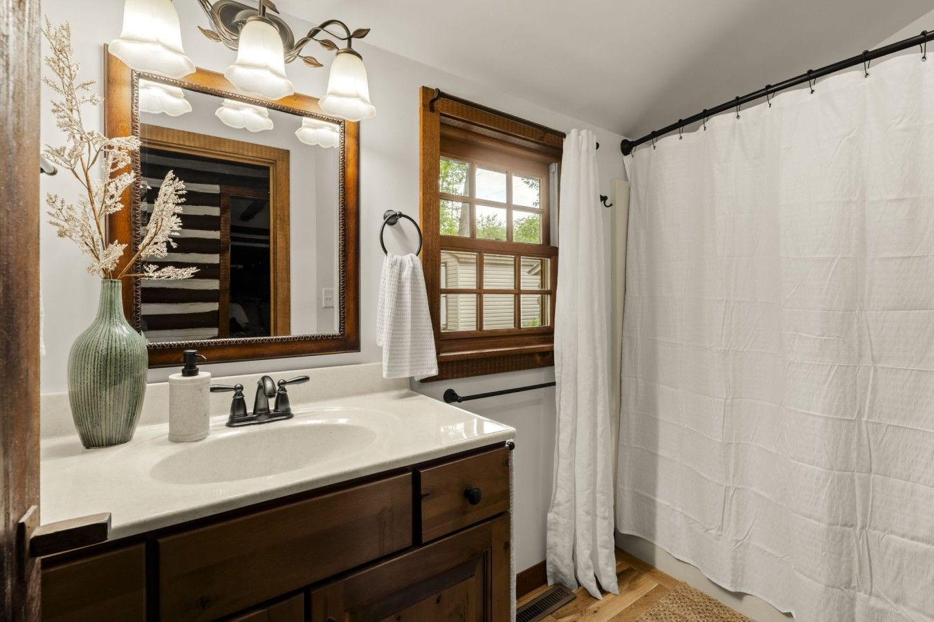 Un baño con tocador de madera, lavamanos blanco, espejo con luces, ventana con vistas y cortina de ducha blanca.