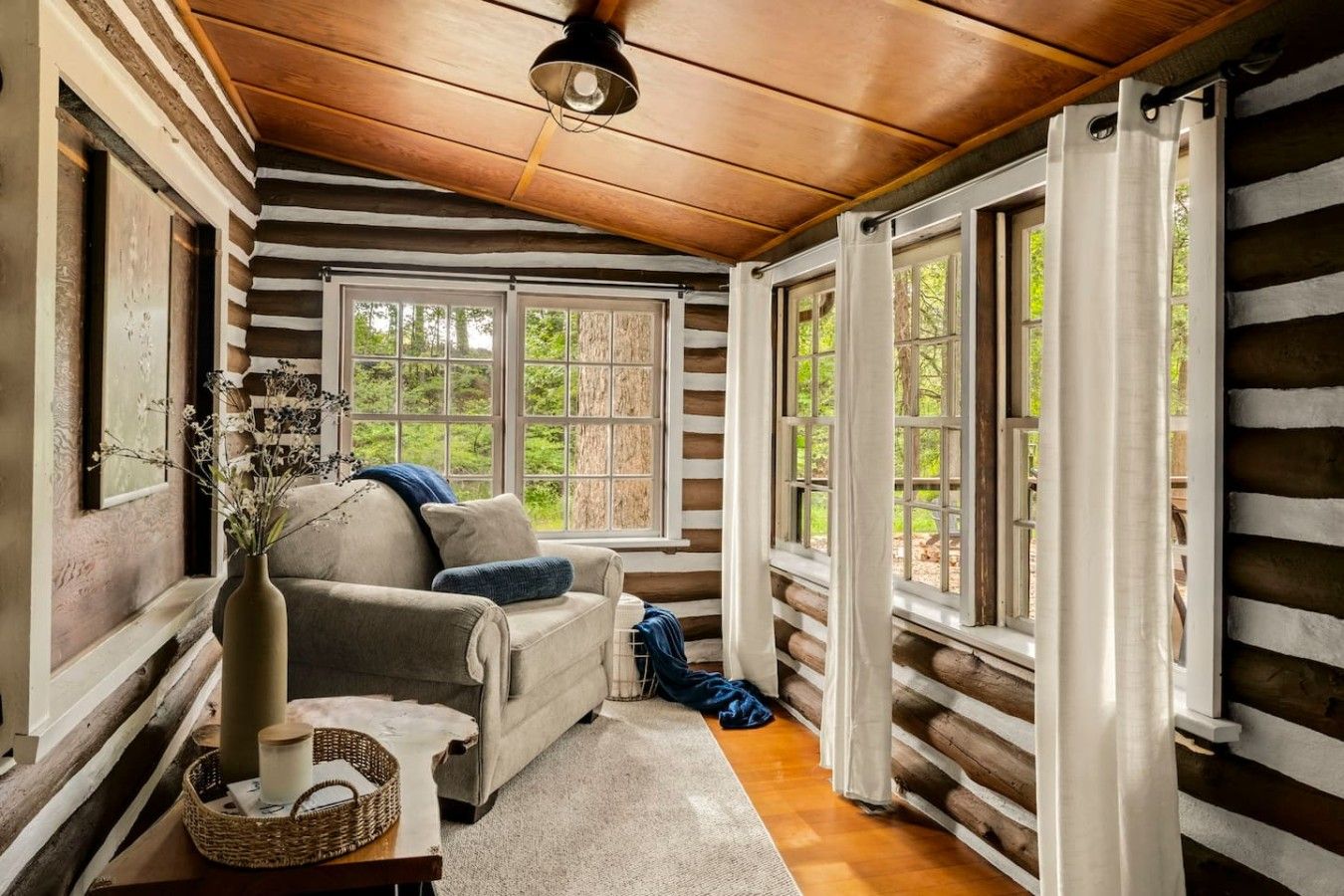 A cozy cabin interior with a wooden ceiling, a sofa, a vase with flowers, and large windows.