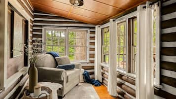 A cozy cabin interior with a wooden ceiling, a sofa, a vase with flowers, and large windows.