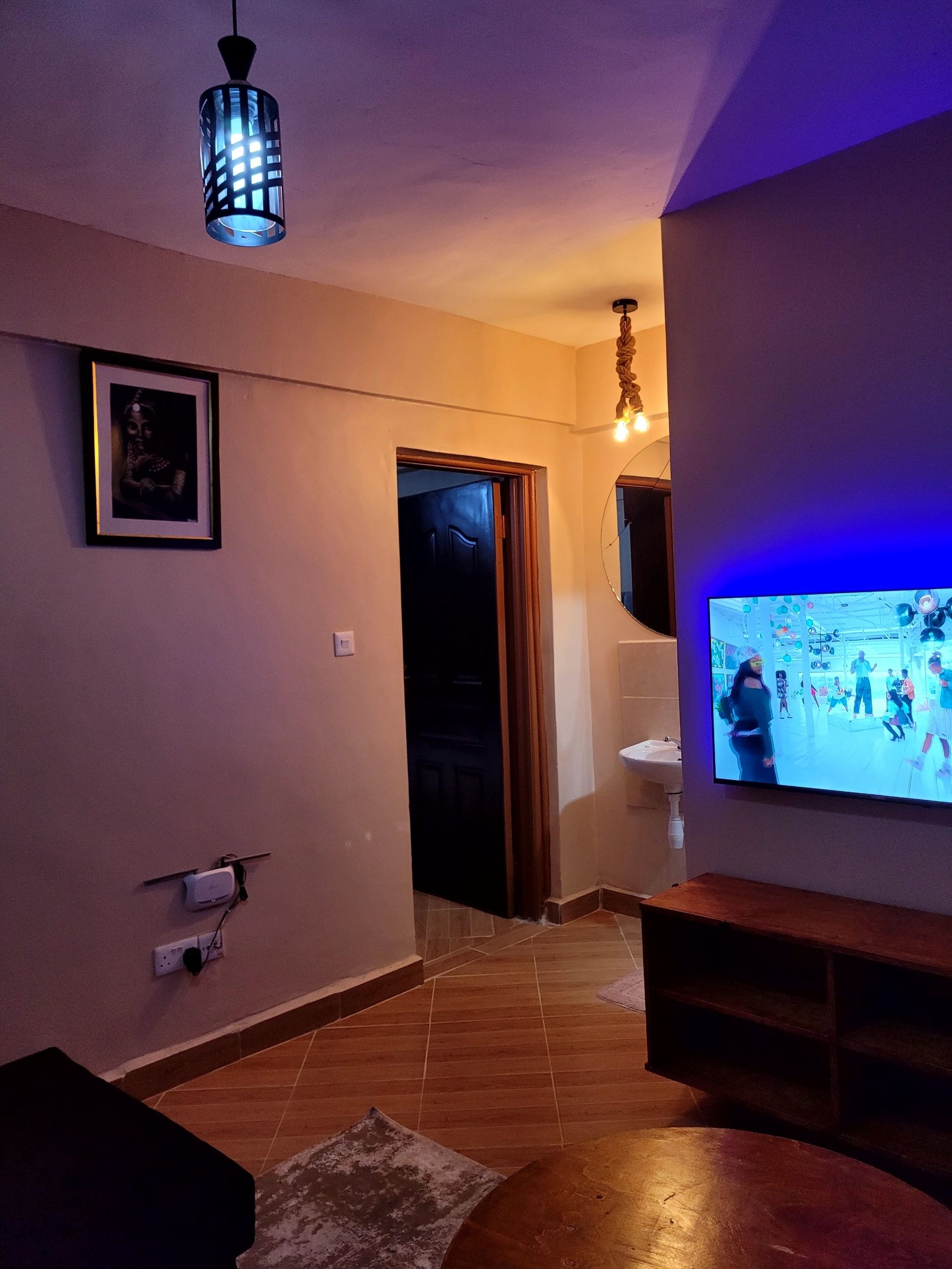 A living room with a television displaying a dance scene, a framed picture on the wall, and a hanging light fixture.