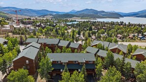 A residential area with multiple two-story buildings, surrounded by dense forest, with a mountain range and a lake in the background. - Dillon 3BR Mountain Townhome | Hot Tub, Sauna, and near Breck, Keystone (Dillon)