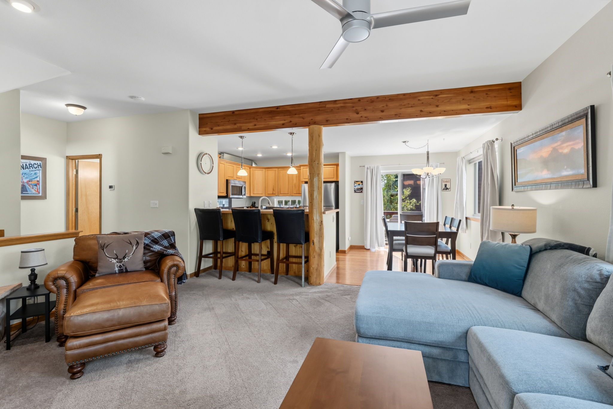 A spacious living area with a sofa, a leather armchair, and a dining table with chairs. A kitchen with wooden cabinets and a refrigerator is visible in the background.