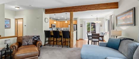 A spacious living area with a sofa, a leather armchair, and a dining table with chairs. A kitchen with wooden cabinets and a refrigerator is visible in the background.