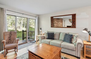 Ein Wohnzimmer mit einer Couch, einem Holztisch, einem Korbstuhl und einem großen Fenster mit Blick auf Grünflächen.