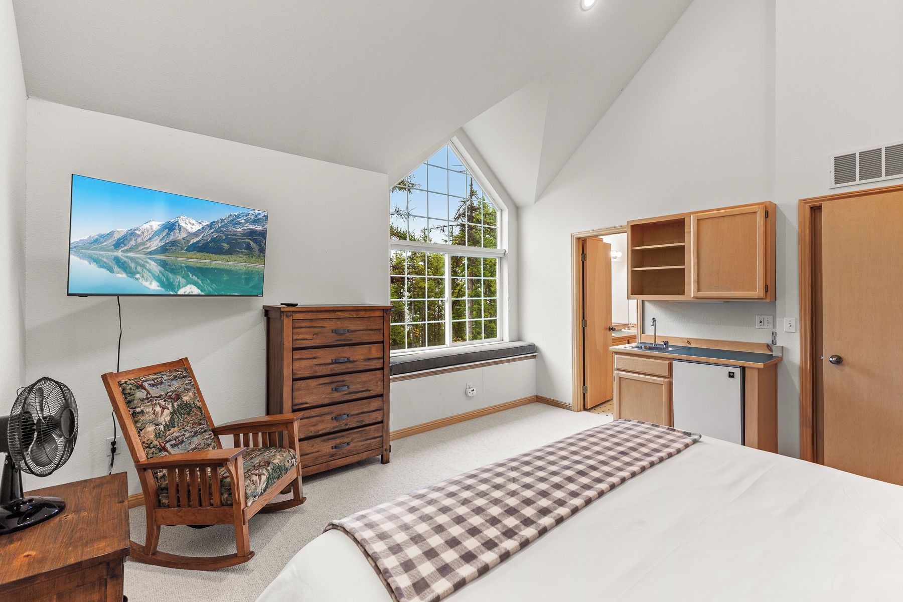 A bedroom with a bed, a dresser, a chair, a desk with a fan, a window with a view, and a kitchenette.