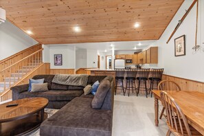 A living room with a sofa, ottoman, and coffee table, a dining area with a wooden table and chairs, and a kitchen with wooden cabinets and a refrigerator. - New! Spacious Ski Retreat | Sleeps 13 | 3 Min to Schweitzer Mountain Resort (Sandpoint)