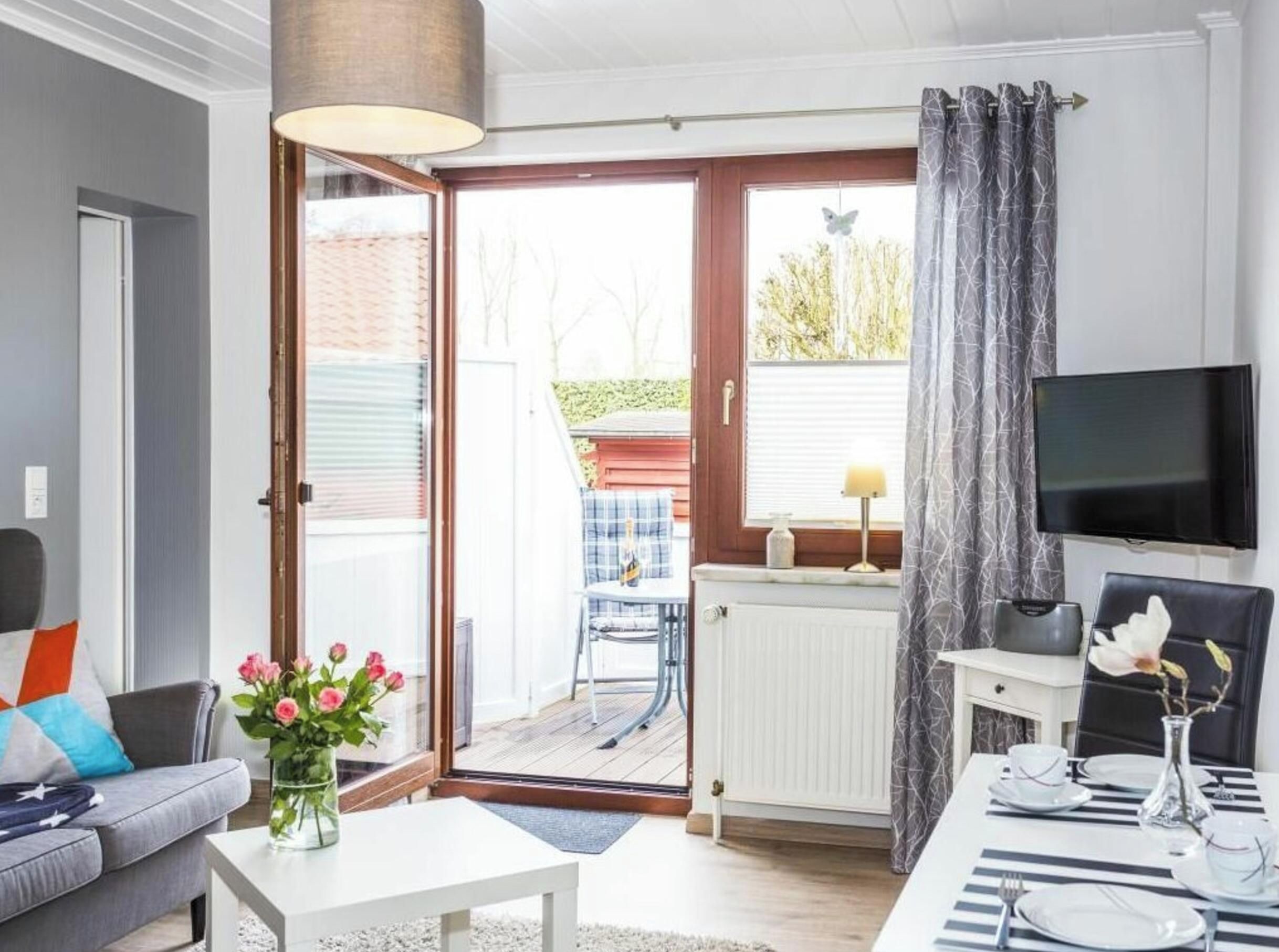 A modern living room with a sofa, a coffee table with flowers, a dining area set for two, and a sliding glass door leading to an outdoor space.