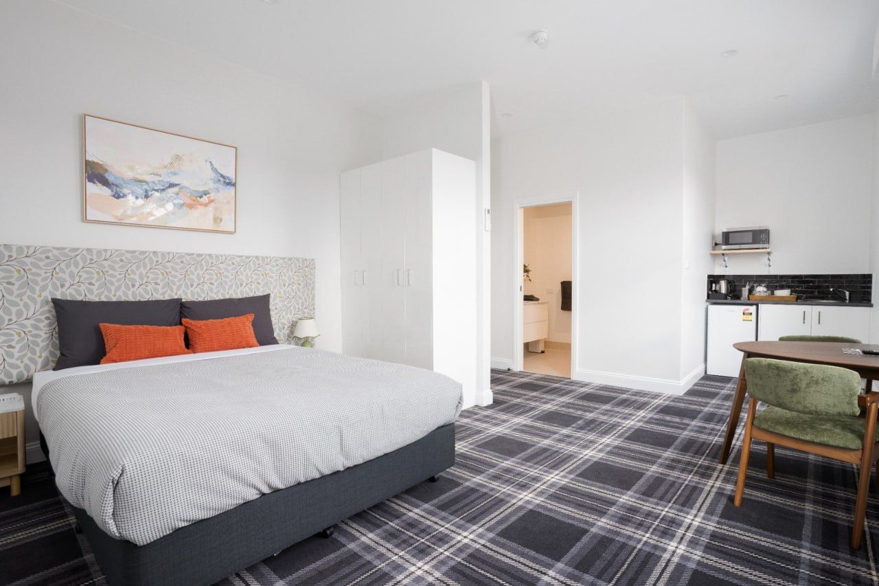 A bedroom with a bed, a nightstand, a bedside table, a green chair, a kitchen area with a microwave, and a door leading to another room.