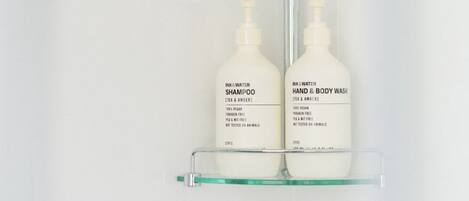 Un baño con cabezal de ducha, un estante de vidrio que sostiene tres botellas de champú, acondicionador y jabón de manos y cuerpo.