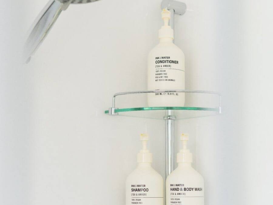 A bathroom with a showerhead, a glass shelf holding three bottles of shampoo, conditioner, and hand & body wash.