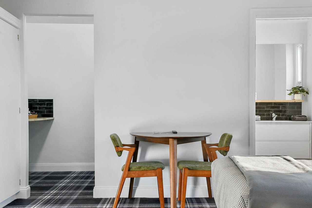 Una habitación con una mesa redonda y dos sillas, una cama con colcha blanca y gris, y una zona de cocina con salpicadero negro.