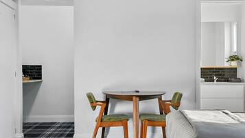 Una habitación con una mesa redonda y dos sillas, una cama con colcha blanca y gris, y una zona de cocina con salpicadero negro.
