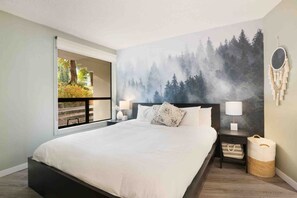 A bedroom with a large bed, a nightstand, a lamp, a wall-mounted mirror, and a view of a balcony with outdoor scenery. - Upper Vill 1BR: Walk to Ski-In/Out, Sleeps 4! Hot Tub, Sauna & Gym. (Whistler)