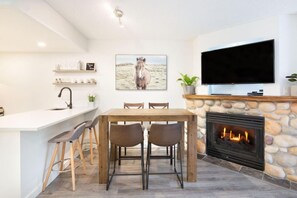 A modern kitchen with a bar area, a dining table with chairs, a fireplace, and a mounted television. - Upper Vill 1BR: Walk to Ski-In/Out, Sleeps 4! Hot Tub, Sauna & Gym. (Whistler)