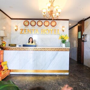Un mostrador de recepción con una araña, relojes y un cartel que dice 'ZENTA HOTEL'.