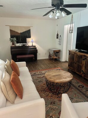 A living room with a white sofa, a round coffee table, a ceiling fan, a television, and a patterned rug. - The Heritage Home, Walk to Downtown (Auburn)