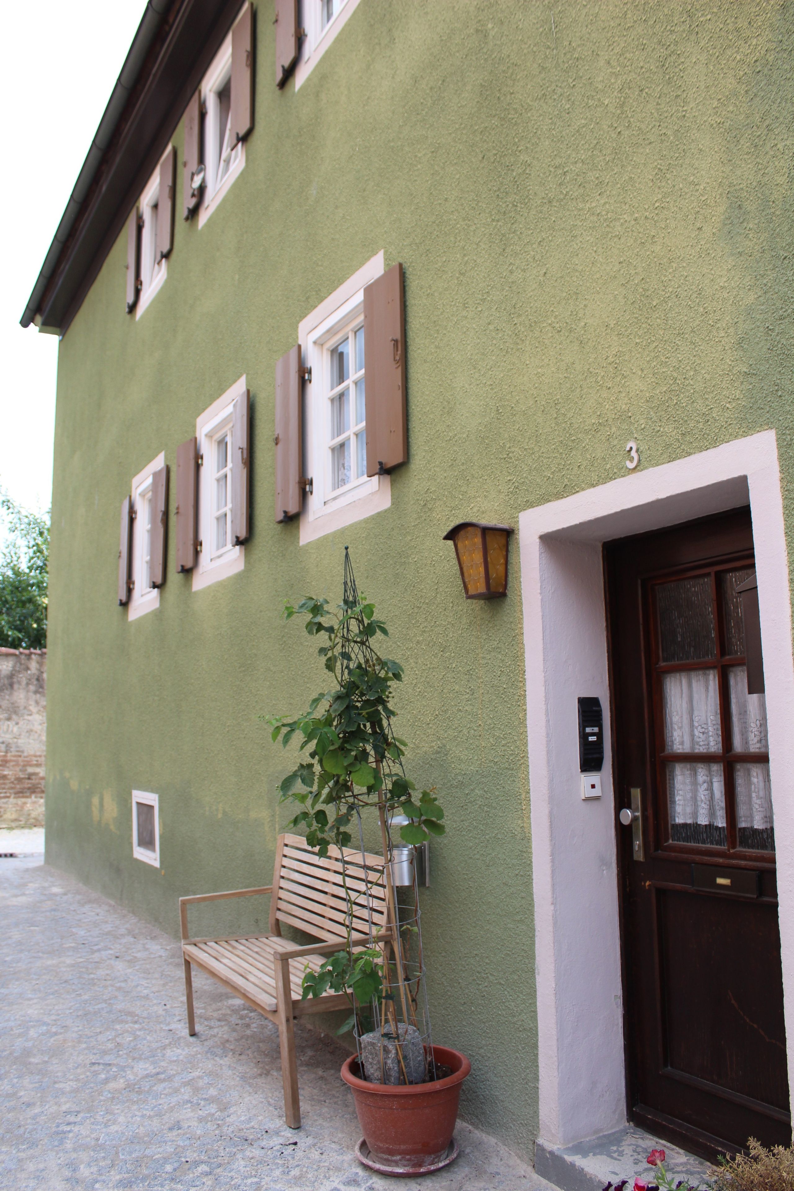 A building with a green exterior, a wooden bench, a potted plant, and a door with a number.