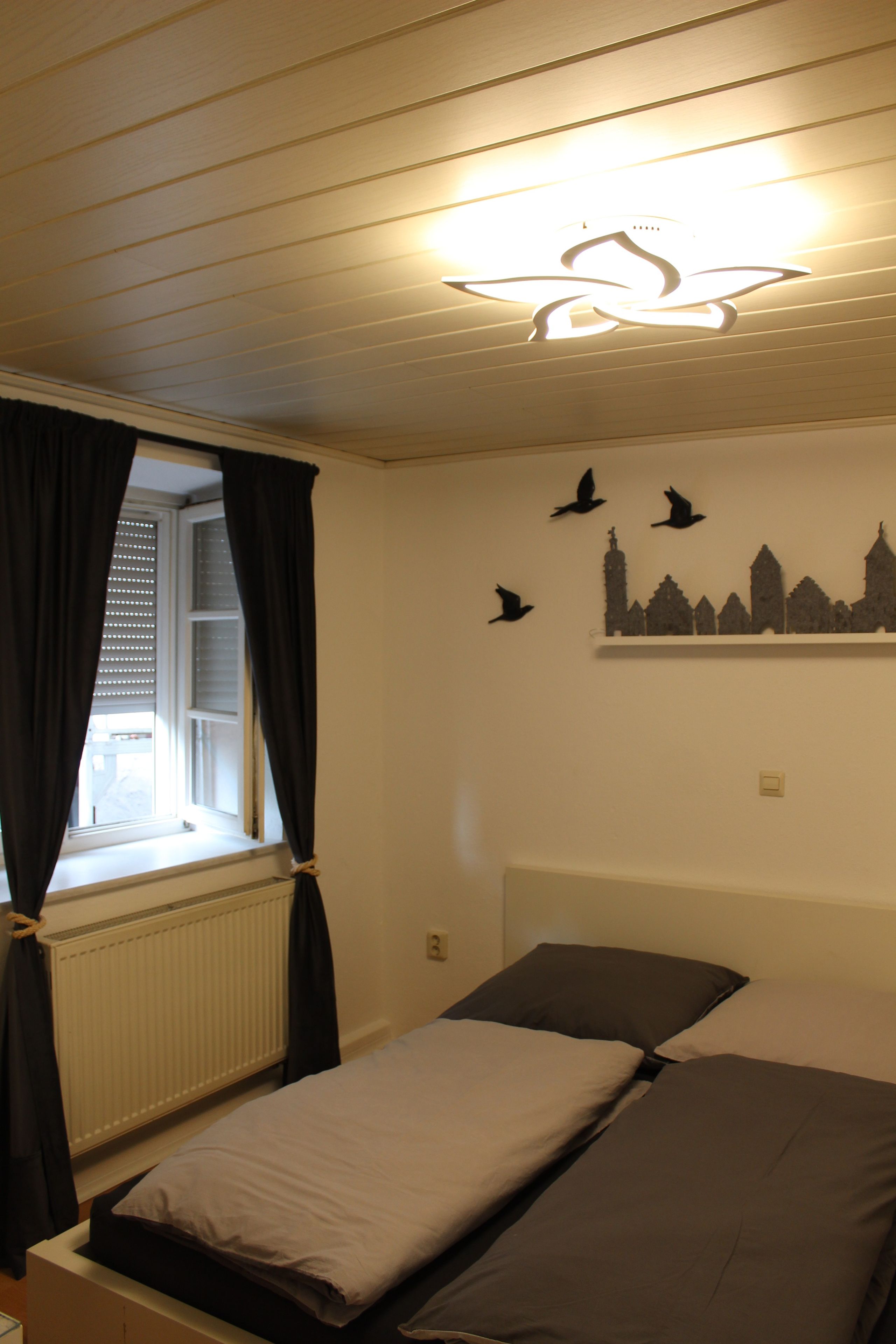 Une chambre à coucher comprenant un lit, une fenêtre avec des rideaux, un radiateur et des éléments de décoration murale, notamment des silhouettes d’oiseaux et une représentation d’un paysage urbain.