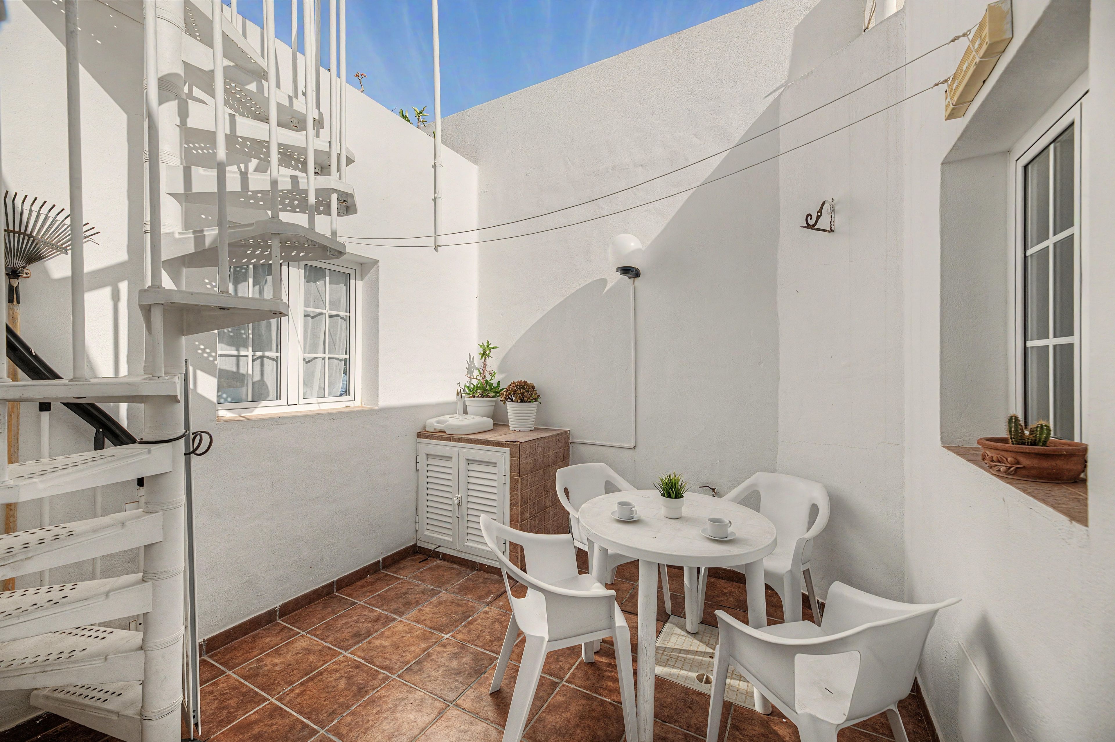 Un pequeño patio cubierto con una mesa y sillas blancas, una planta en maceta y un ventanal.