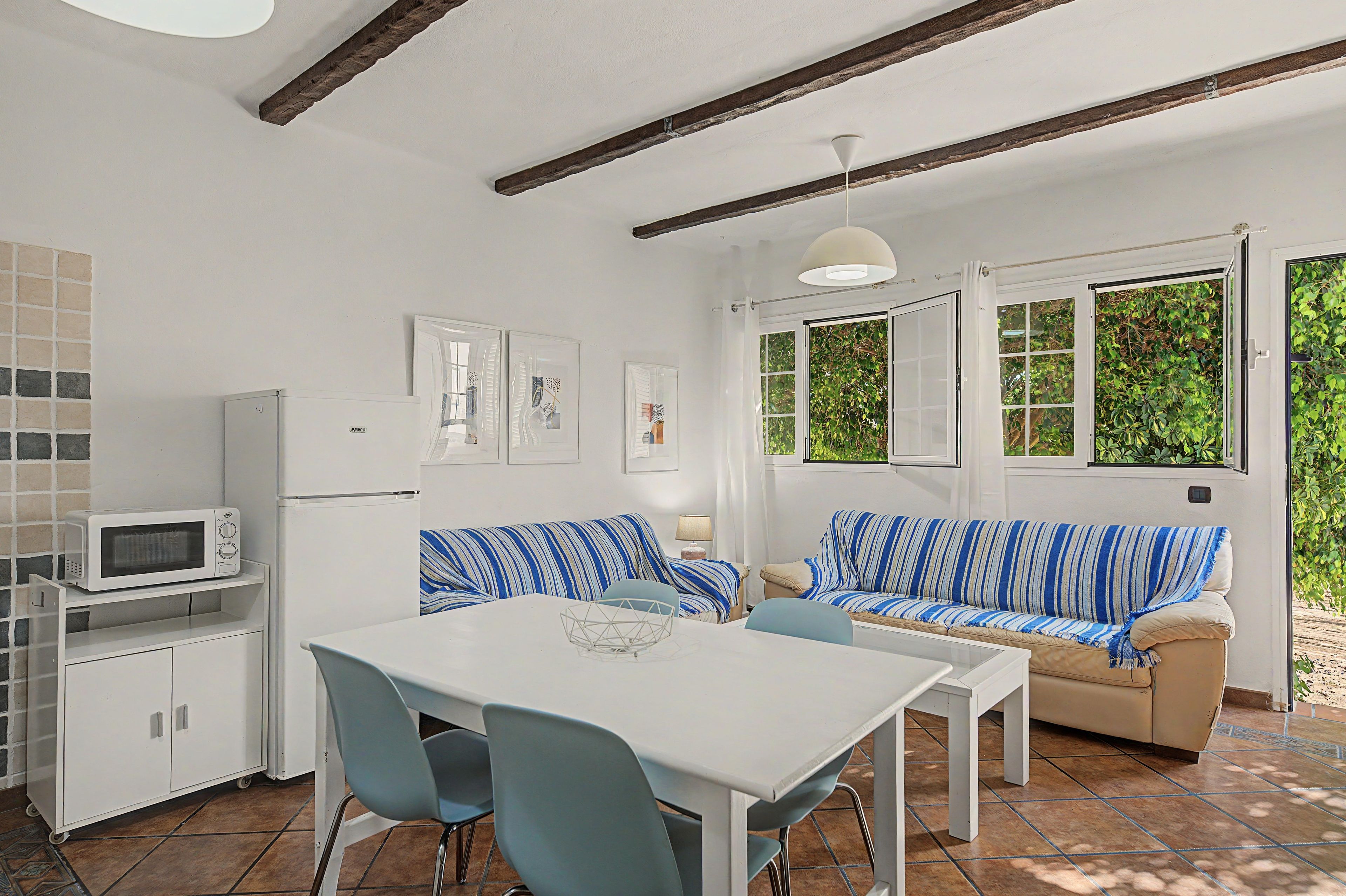 A compact kitchen with a microwave, a refrigerator, and a dining area with a table and chairs. There is a sofa with striped cushions and a window with a view of greenery outside.