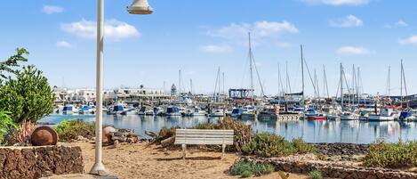 Ein sandiger Strand mit einer Bank und einer Straßenlaterne, mit Blick auf einen Yachthafen mit zahlreichen Booten.