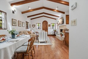 A dining area with a long table set for a meal, wooden beams on the ceiling, and a fireplace with a stone surround. - 4090 Oceans of Time - Stunning Carmel Cottage (Carmel-by-the-Sea)