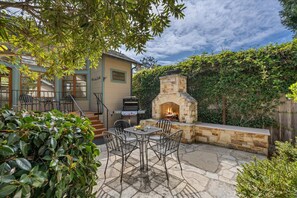 Un patio exterior con un hogar de piedra, una parrilla y un comedor con sillas y mesa de metal.