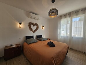 A bedroom with a bed, a nightstand, a wall-mounted heater, a heart-shaped decoration, and a window with curtains. - Villa Victoria 5*, Oceanfront, Pool, Air Conditioning, Renovated Home, New Amenities (L'Aiguillon-la-Presqu'île)