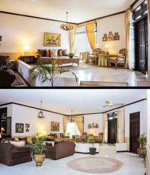 A living room with a sofa set, a coffee table, and a chandelier. There is a dining area with a table and chairs. The room has a large window with curtains.