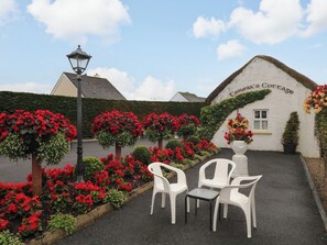 A cottage with a thatched roof, surrounded by a garden with red flowers and a paved area with white plastic chairs and a table. - TERESA'S COTTAGE, pet friendly in Bruckless, County Donegal (BRUCKLESS)