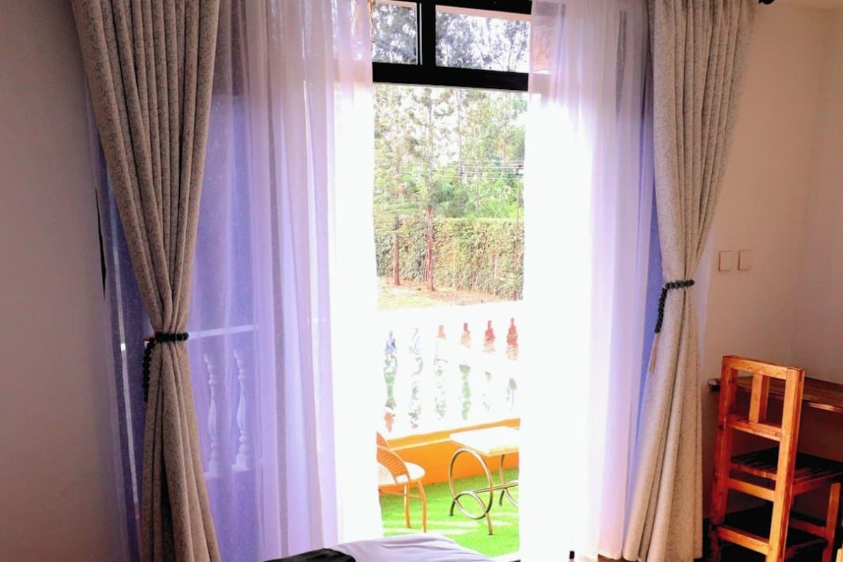 A neatly made bed with a towel, a wooden chair, and a view of an outdoor area through a window with curtains.