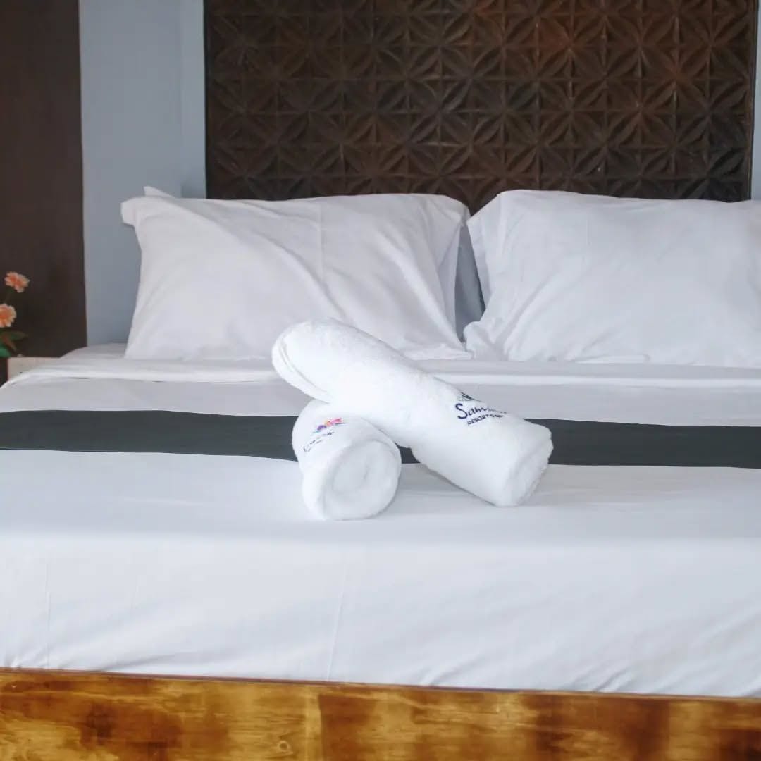 Una cama bien hecha con sábanas y almohadas blancas, una toalla enrollada y un cabecero de madera con un diseño geométrico.