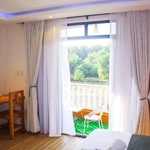 A hotel room with a bed, a wooden chair, a small table, and a balcony with a view of greenery.
