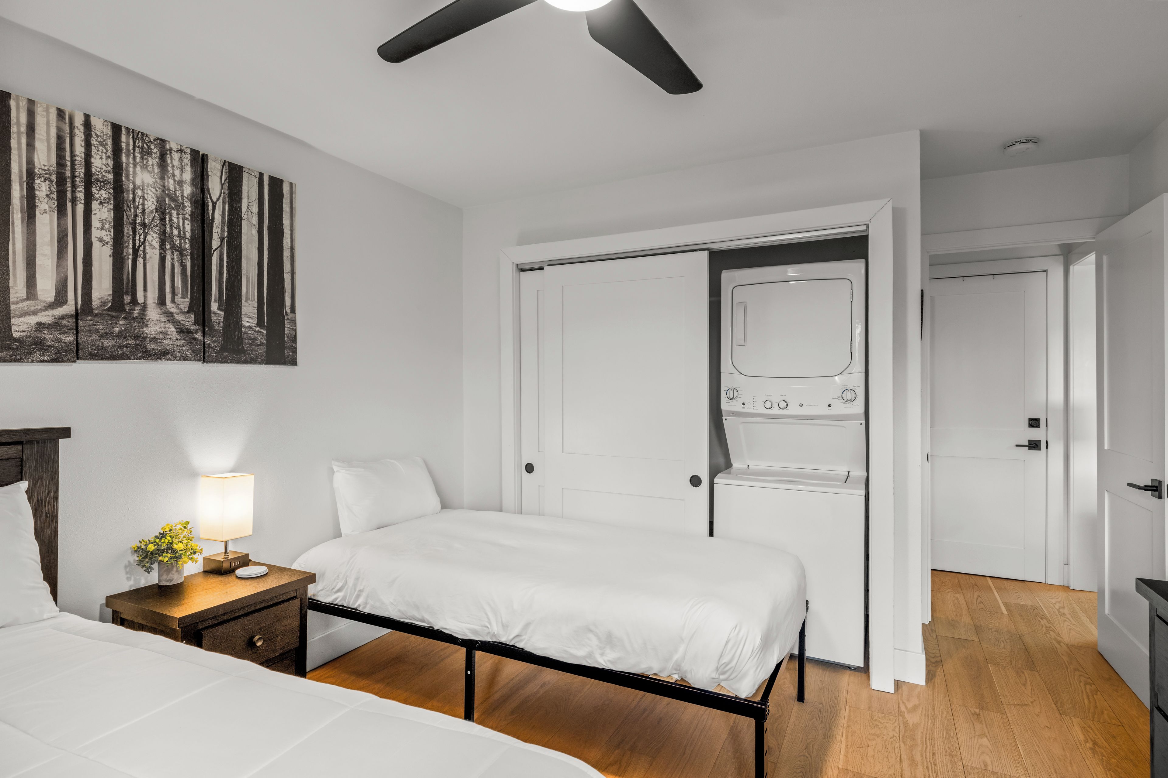 Quarto de hotel com cama, mesa de cabeceira, luminária e uma pintura de uma floresta.